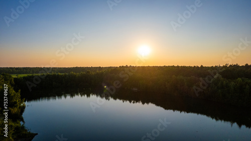 This is an aerial landscape photograph capturing a tranquil scene at sunset. The sun is positioned low in the sky, casting a warm golden glow over the scenery. It hangs just above a dense forest that