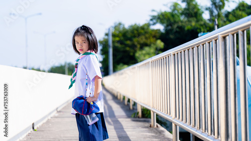 A schoolgirl walks home from school in the hot summer sun, a girl frowns against the hot sun, Child walks on an overpass, looks back at the camera, a 6 year old Kid wears a Thai school uniform. © Kanthita