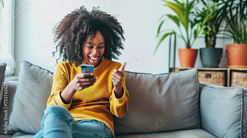 Happy young black woman sitting on sofa at home with phone in her hand. photo