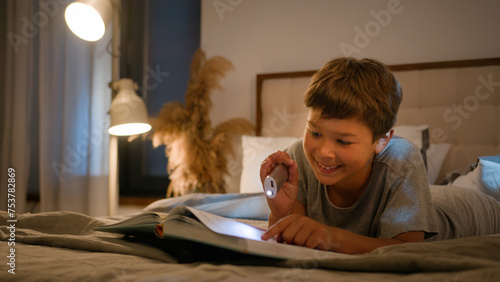 Surprised amazed smiling funny Caucasian child kid boy schoolboy reading interesting book evening in bedroom using flashlight lying on bed home enjoying read fairytale amazing story for night bedtime