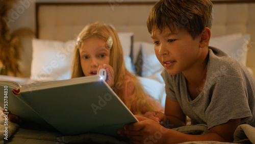 Caucasian children kids boy girl older brother younger sister lying on bed at home evening reading interesting book using flashlight read fairytale story for night bedtime happy family relationships