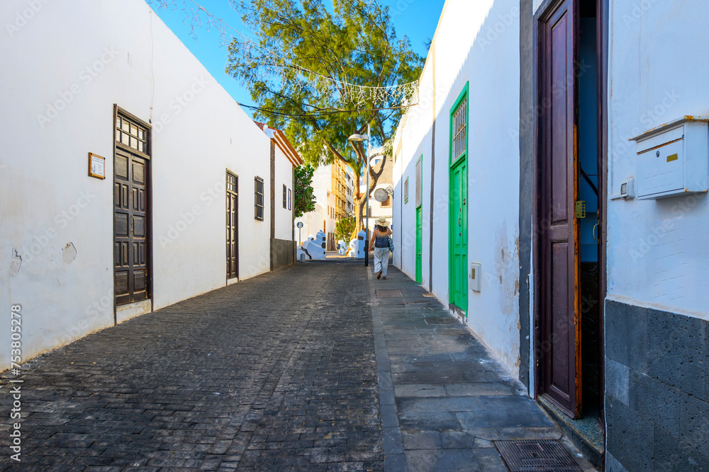 Obraz premium A pedestrian walks down a picturesque street of whitewashed homes with colorful green, wooden, and turquoise doors in the old town center of Arrecife, Spain, on the Canary island of Lanzarote.