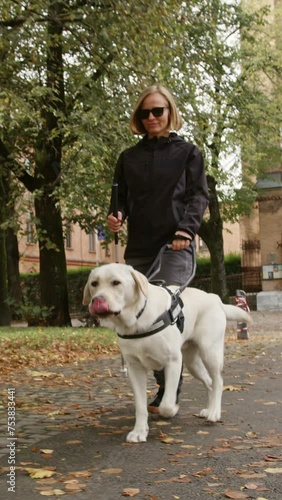 Blind woman in the company of her loyal guide dog safely and relaxed walking along a city park, front view vertical handheld shot.