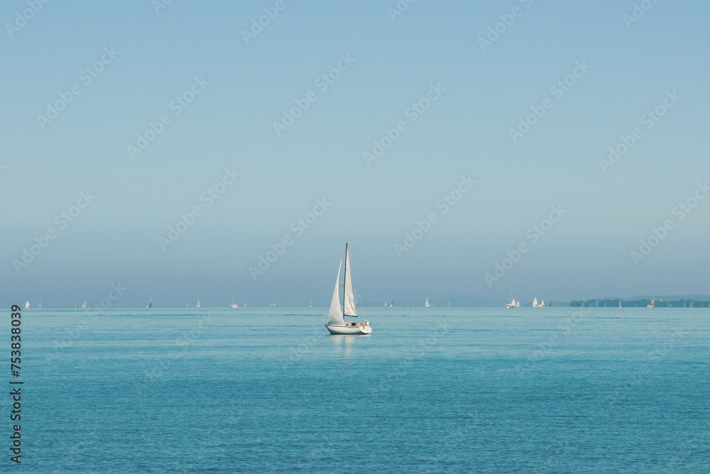 Small white sailboat in blue lake with many other boats far in background. Sailing yacht with one mast on a sunny day in big lake. Blue water and blue sky. Calm water without waves.