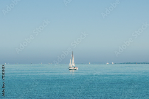 Small white sailboat in blue lake with many other boats far in background. Sailing yacht with one mast on a sunny day in big lake. Blue water and blue sky. Calm water without waves.