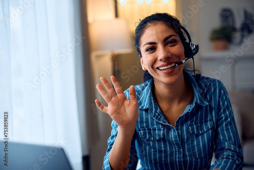 Happy Latin entrepreneur waving while working at her home office and looking at camera.
