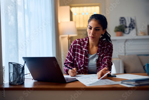 Latin American woman using laptop while working on documents at home.