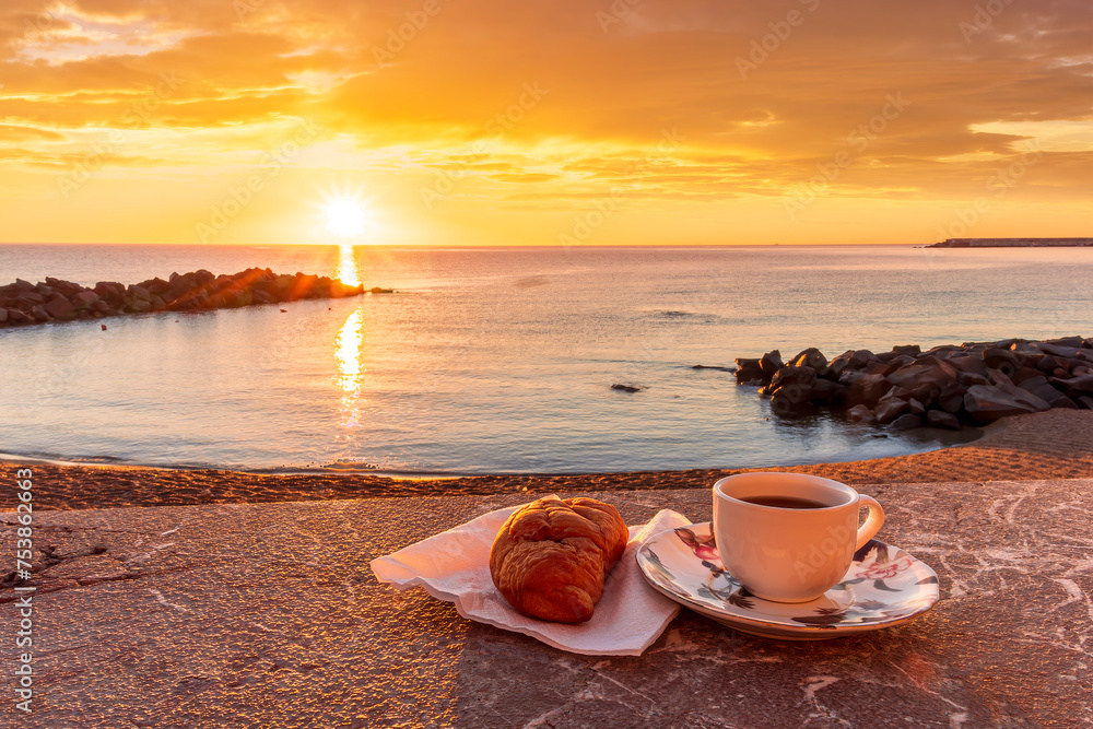cup of coffee or tea with croissant on a morning embarkment beach with blue sea and beautiful cloudy sunrise or sunset on background, street food breakfast concept