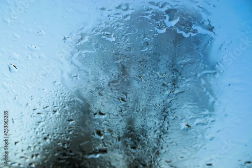Abstraktes Motiv mit Eis- und Wassermuster auf beschlagener Glasscheibe vor hohem Baum und blauem Himmel mit Licht bei Frost, Kälte und Sonne am Morgen im Winter