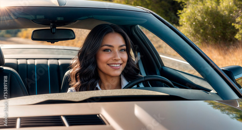 A woman with long dark hair is sitting behind the wheel of a car. She is smiling and looking at the camera.