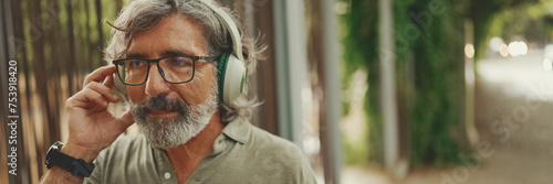 Panorama of friendly middle-aged man with gray hair and beard wearing casual clothes listening to music on headphones. Mature gentleman in eyeglasses enjoys music outdoors