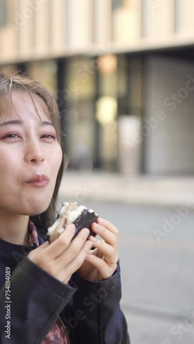 A vertical video of a young Japanese Okinawan woman in her 20s eating fast food pork egg rice balls on Kokusai Street, a tourist attraction in Naha City, Okinawa Prefecture 沖縄県那覇市の観光名所の国際通りで若い２０代の日本人沖 photo