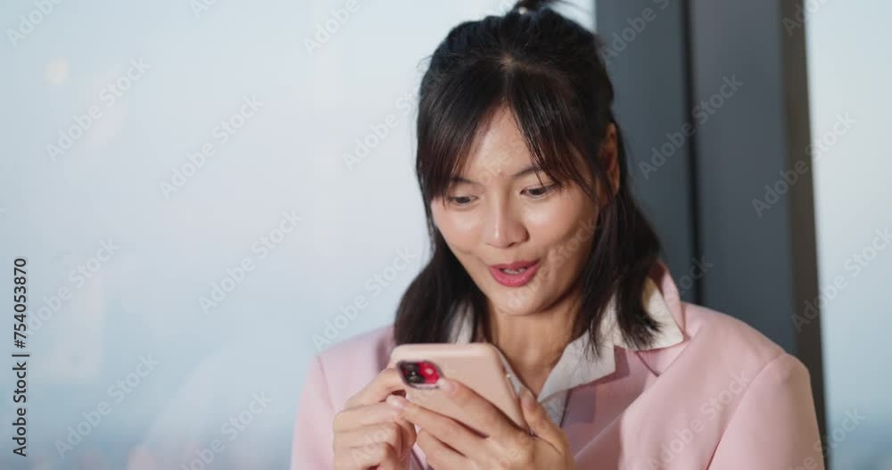 A delighted young woman in a pink coat reacts with surprise while looking at her smartphone screen inside a bright room. A delighted young woman in a pink coat reacts with surprise while looking at her smartphone screen inside a bright room.