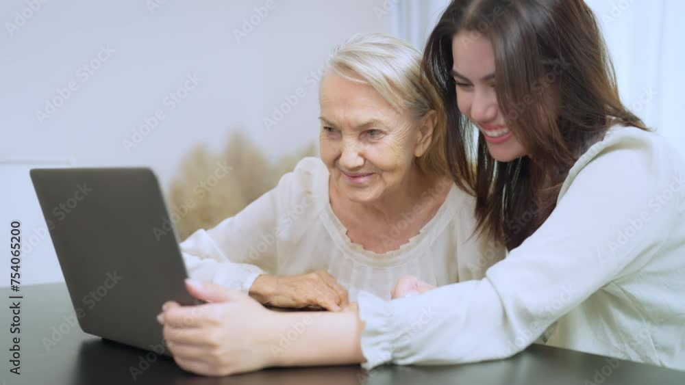 Cheerful young woman guides an elderly woman through laptop use, a display of patience and intergenerational learning. Cheerful young woman guides an elderly woman through laptop use, a display of patience and intergenerational learning.