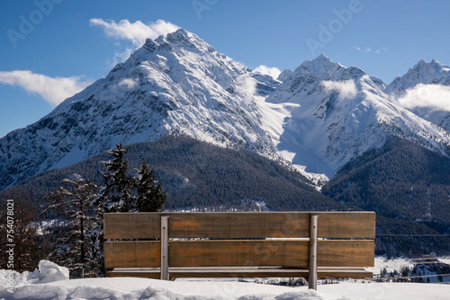 Alpine mountain panorama in winter.  The wooden backrest of a beautifully situated bench in the Engadin, Switzerland.  Snow-capped mountain peaks and white clouds against a blue sky photo