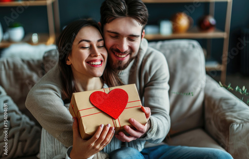 Happy young couple with gift box and red heart shaped paper on Valentine's Day at home
