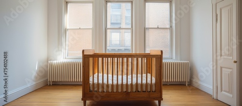 A simple wooden baby crib sits in a white room of a contemporary apartment, illuminated by natural light streaming in through a window. The room is small, with a door visible in the background.