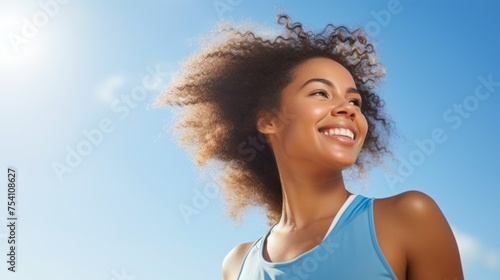 A happy African American female athlete smiles while jogging  exercising  exercising outdoors against a blue sky background. Summer  Sports  Fitness  Running  Healthy Lifestyle concepts.