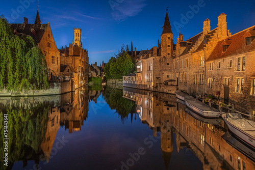 Bruges canal in the evening. Rosary Quay in the center of the old town of the Hanseatic city. Illuminated historic merchant houses with reflections on the water surface. Boats at the jetty