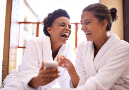Beauty, phone and spa with women laughing in robes for luxury pampering or treatment together. Happy, app and social media with funny young friends at resort or salon for wellness or weekend getaway