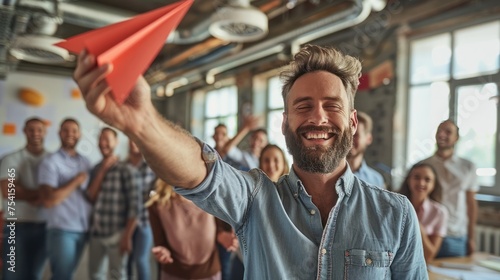 Happy male leader of company throwing red paper plane in office with group of employees smiling and talking in background.