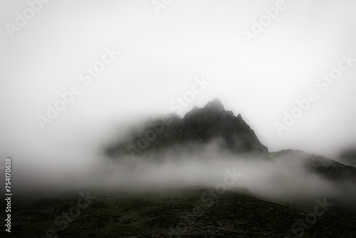 Overcast alpine view with mountain silhouette fragments through fog and clouds. Climate, environment and weather concept sky background