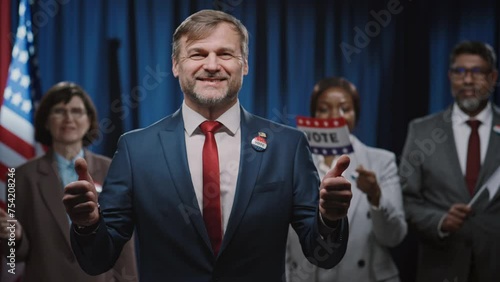 Medium portrait shot of cheerful Caucasian male candidate for presidency, Senate or Congress, showing thumbs up, smiling, and multiethnic team waving vote flags and cheering photo