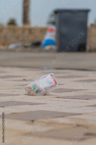 A bag of garbage is lying on the sidewalk, signaling that it was not thrown to the trash can in the background nearby or flew out of it during shipment, the lack of a lid, the wind blew away