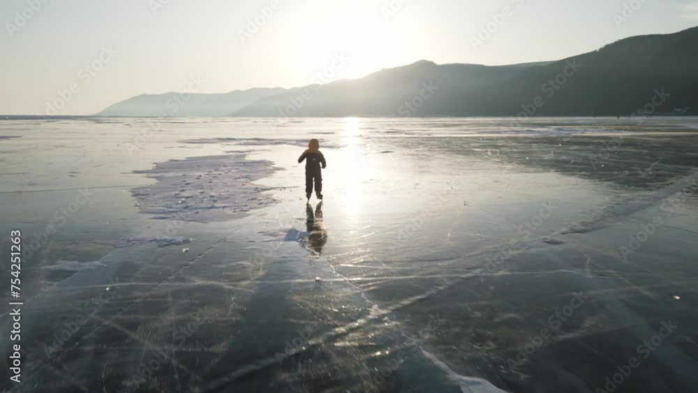 A child in a down jacket is clumsily skating on the blue transparent ice of the endless winter Lake Baikal at sunset A child in a down jacket is clumsily skating on the blue transparent ice of the endless winter Lake Baikal at sunset