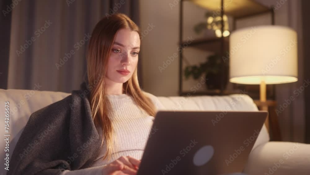 Beautiful young woman relaxing at home or working online browsing products in internet store reading news checking email or social media profile on laptop computer sitting on couch at night indoors Beautiful young woman relaxing at home or working online browsing products in internet store reading news checking email or social media profile on laptop computer sitting on couch at night indoors
