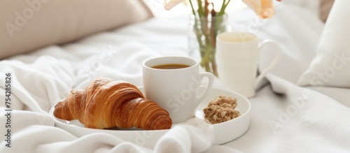 A closeup shot of a white luxury bed with a tray holding two mugs of coffee, croissants, and an orchid flower. The scene depicts a breakfast setup in a cozy setting. photo