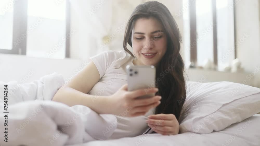 Charming smiling young woman hold smartphone scrolling app watching social media feed laying on bed indoors Happy female texting laughing enjoying leisure time at cozy home Charming smiling young woman hold smartphone scrolling app watching social media feed laying on bed indoors Happy female texting laughing enjoying leisure time at cozy home