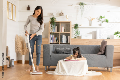 Asian Mother vacuuming the floor while a little girl focuses on drawing at home