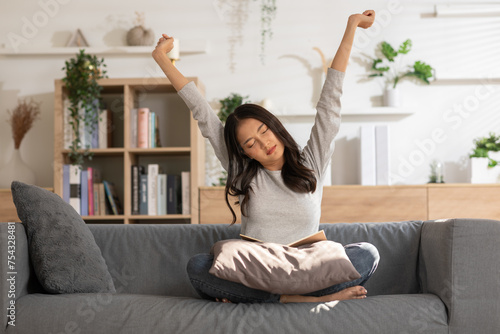 Relaxed young woman stretching with closed eyes while sitting with a book on a cozy sofa