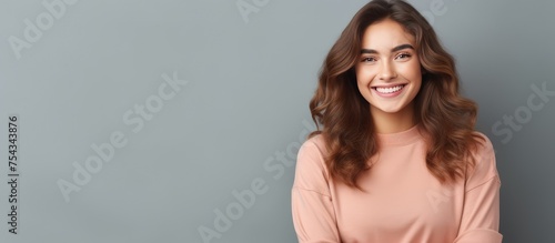 A joyful European student, laughing at a good joke, stands in front of a vibrant blue wall. She keeps her hands crossed and wears casual clothes with round spectacles. The happy young woman with long