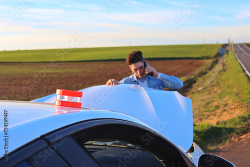 Caucasian man calling on the phone looking at the engine with the emergency light V16, daytime. Concept of problem on road