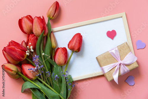 Red tulips with gift box and small hearts on pastel pink background with empty space.
