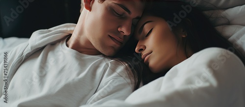 A beautiful young couple, wearing the same white hoodie and black pants, is laying in bed together. The man gently hugs his beloved woman from behind while they both dream of a happy future and enjoy photo