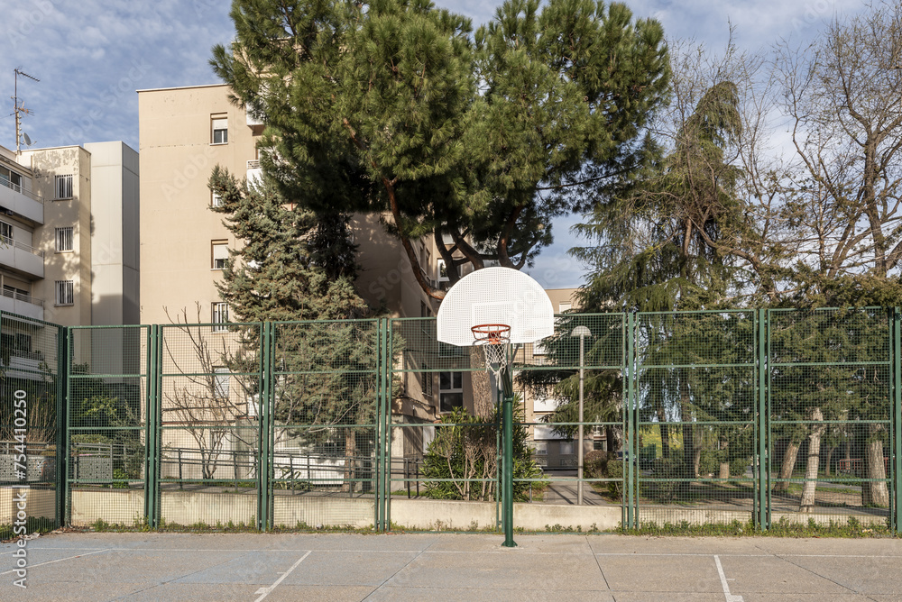 An urban basketball court with rough cement floors, a perimeter with metal fences and baskets on metal posts screwed to the ground in an area with trees and buildings An urban basketball court with rough cement floors, a perimeter with metal fences and baskets on metal posts screwed to the ground in an area with trees and buildings