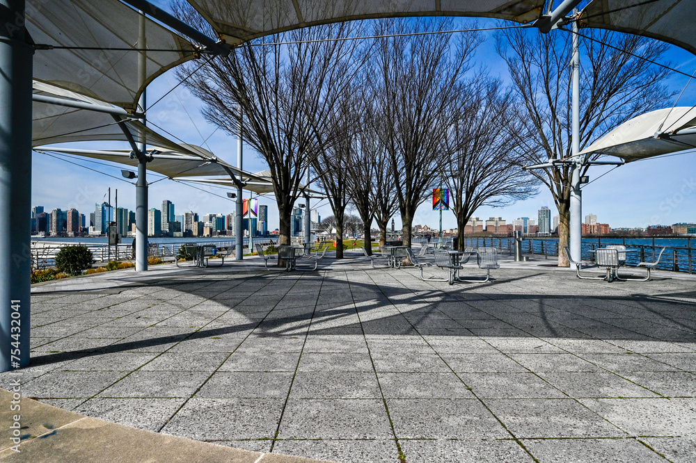 Scenic view of NBC West Street with outdoor seating under white canopies, overlooking the tranquil Hudson River and distant cityscape.
