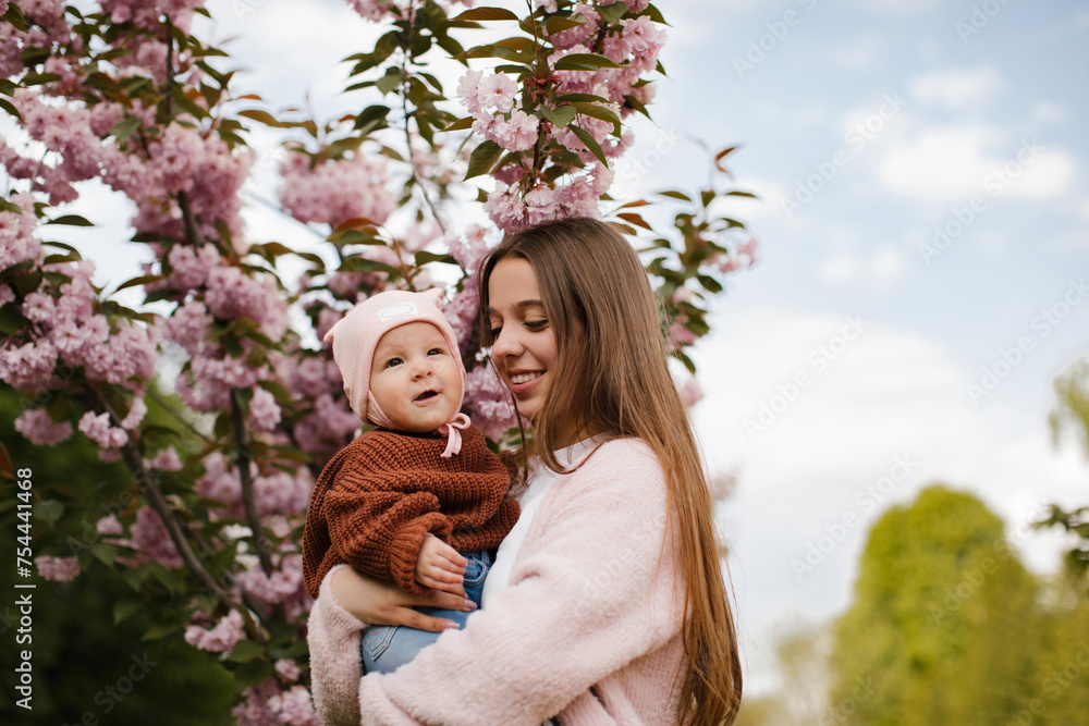 Obraz premium A teenage girl with long hair in a pink jacket is holding a small child near a blooming cherry blossom. The older sister is holding his little sister and smiling near the cherry blossom. 