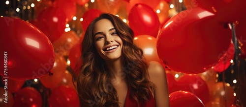 A beautiful woman is standing in front of a bunch of red balloons, smiling and dancing at a Christmas party. She is celebrating the New Years Eve with colorful decorations around her. © TheWaterMeloonProjec
