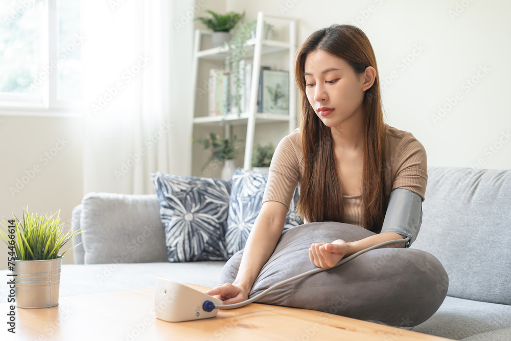Hypotension, asian young woman sitting on couch checking blood high pressure and heart rate with digital pressure monitor machine making self check up on her arm at home. Healthcare and medical. Hypotension, asian young woman sitting on couch checking blood high pressure and heart rate with digital pressure monitor machine making self check up on her arm at home. Healthcare and medical.