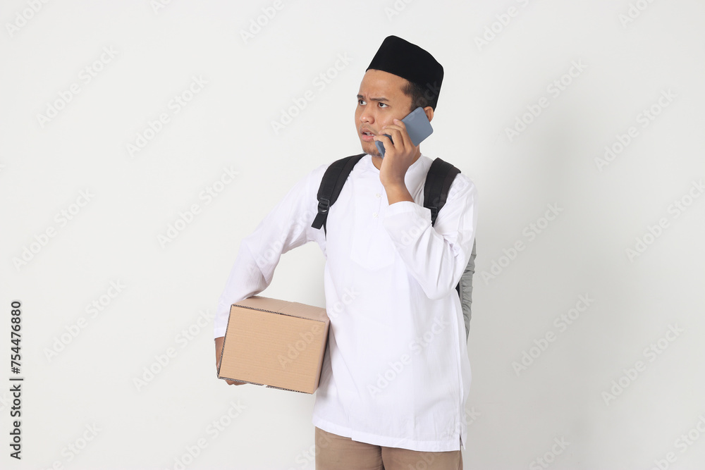 Portrait of worried Asian muslim carrying cardboard box while having having call on mobile phone, showing sad expression. Going home for Eid Mubarak. Isolated image on white background