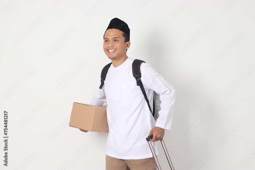 Portrait of excited Asian muslim man in koko shirt with peci carrying cardboard box and holding suitcase handle. Going home for Eid Mubarak. Isolated image on white background