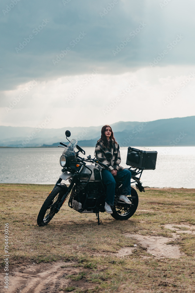 Obraz premium A serene moment captured as a Latina woman sits atop her motorcycle, enjoying the lakeside view with mountains in the distance