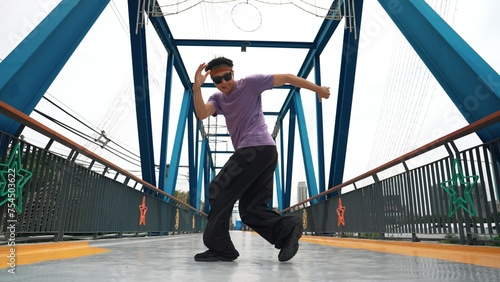 Hipster perform b-boy footstep and street dance at bridge with background. Professional break dancer wear stylish cloth and looking at camera while pose at camera. Outdoor sport 2024. Sprightly.
