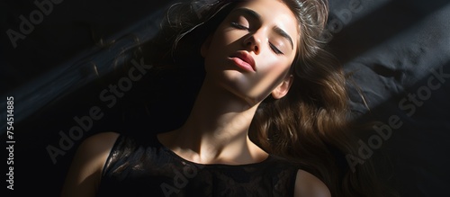 An attractive teenage girl in a black dress is laying down with her eyes closed, her face partially covered by a shadow, in a serene and peaceful pose.