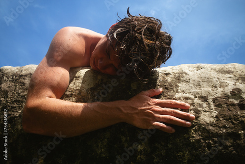 A shirtless young man lies with his head and left arm hugging the surface of a boulder above water in the sun photo