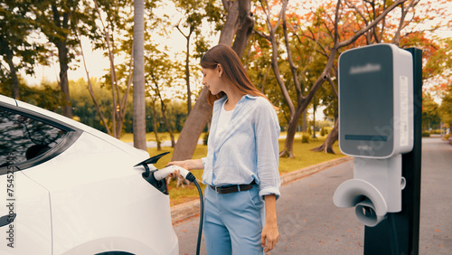 Woman recharging battery for electric car during road trip travel EV car in autumanl season natural forest or national park. Eco friendly travel during vacation and holiday. Exalt photo